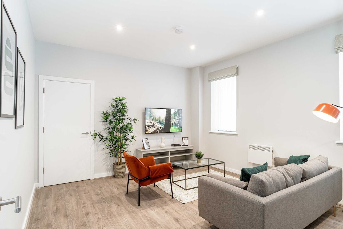 A spacious living area is presented with a comfortable gray sofa and an orange armchair. A low coffee table is centered on a light area rug. A flat-screen TV is mounted on the wall across from the seating. Natural light enters through large windows, complemented by neutral decor.