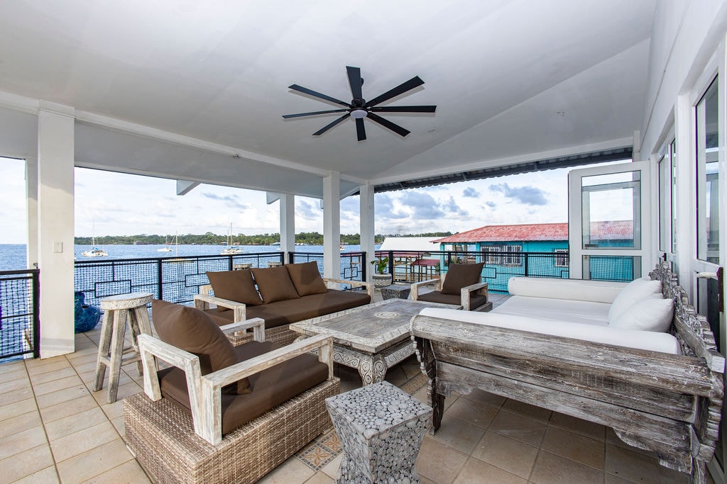 An expansive covered balcony offers several seating areas, featuring brown cushioned sofas and a custom wooden coffee table. A ceiling fan hangs above, providing a cooling breeze. Scenic water views are visible beyond the railing, enhancing the outdoor living experience.