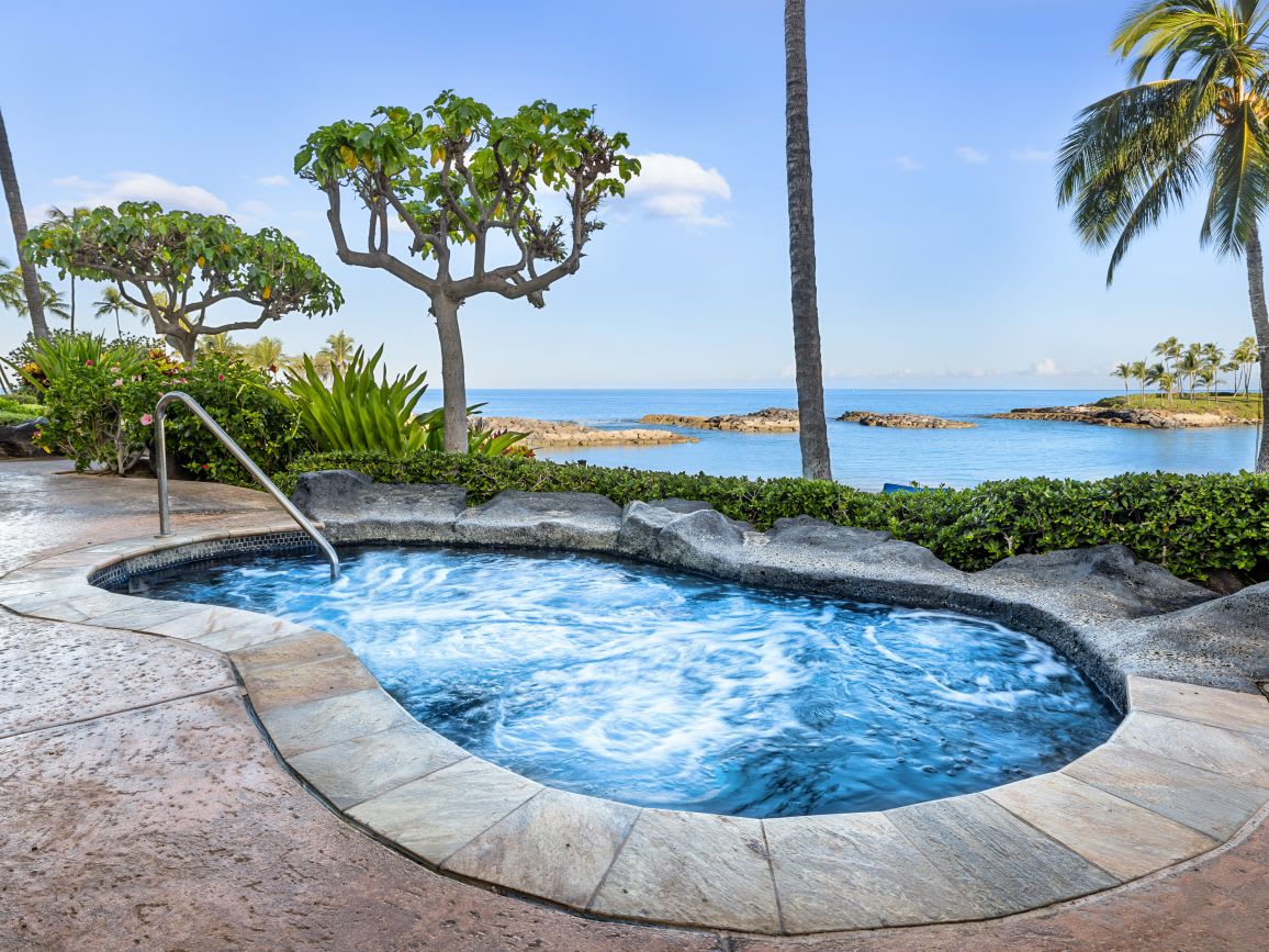 A private hot tub is surrounded by lush greenery and palm trees, offering an unobstructed view of the ocean. The water is bubbling gently, creating a relaxing atmosphere. The stone surround complements the natural landscape, enhancing the sense of tranquility.