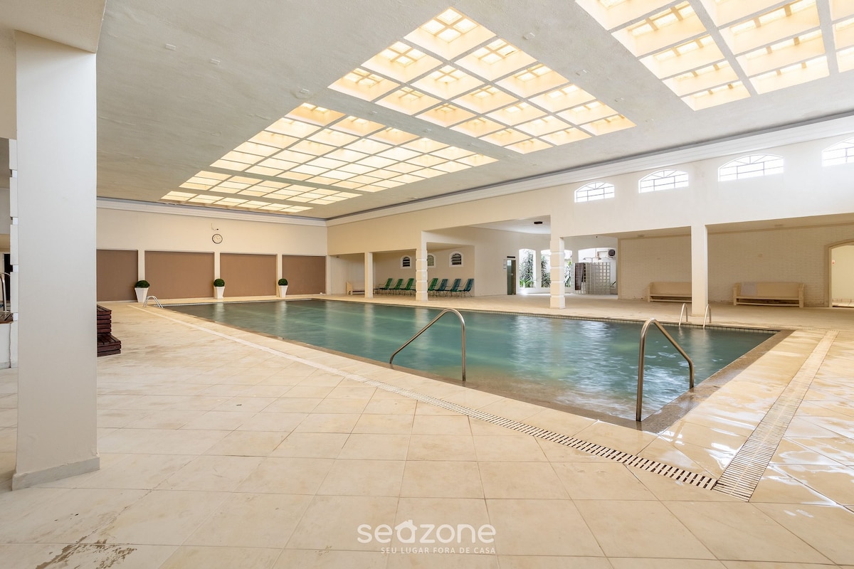 An indoor swimming pool is featured, surrounded by white tiled flooring and walls. Natural light filters through a ceiling with a grid design, creating a bright atmosphere. Poolside loungers can be observed along one side, enhancing the space for relaxation.