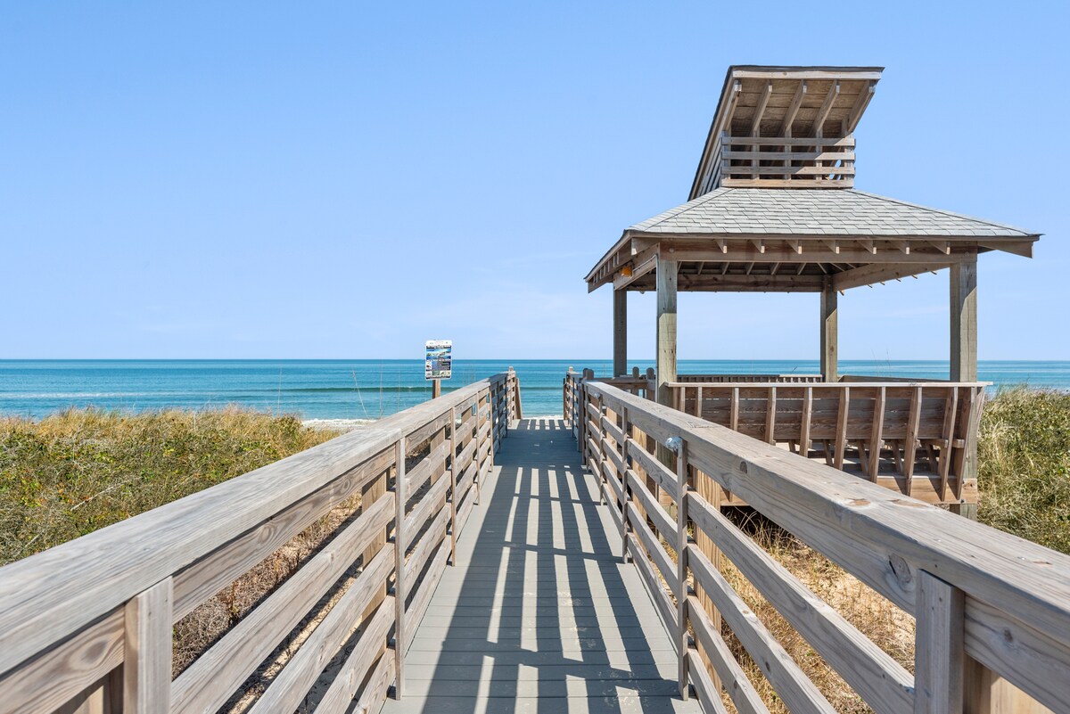 A wooden walkway leads to a gazebo overlooking the ocean. The structure features an open design with a slanted roof, providing an area for shade. Lush greenery borders the path, with expansive ocean views visible in the background under a clear blue sky.