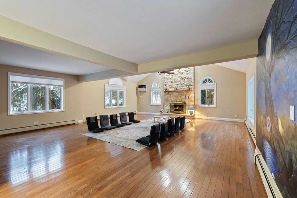 A spacious living area features warm wooden floors and natural light streaming through multiple windows. A stone fireplace serves as a focal point, surrounded by a large area rug. Floor cushions are arranged in a welcoming circle for gatherings or activities.