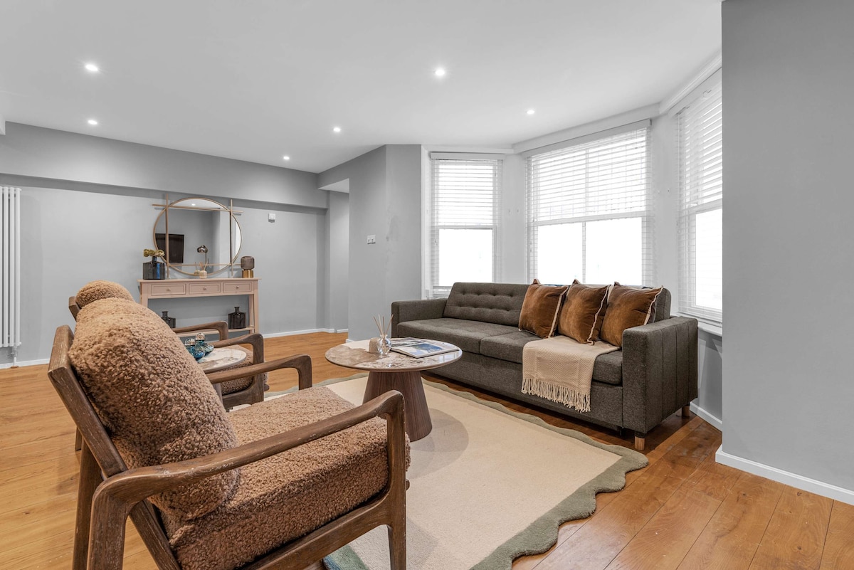 A bright living area is designed with large windows, allowing natural light to fill the space. A comfortable sofa is complemented by plush cushions, while two textured armchairs flank a round coffee table. A decorative mirror and a console table add functionality to this inviting room.
