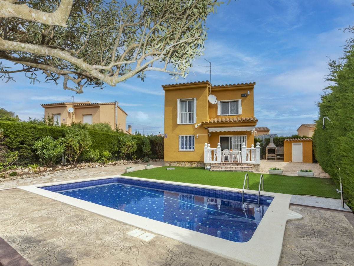 A two-story villa is surrounded by a well-maintained garden. The exterior features yellow walls, white accents, and multiple windows. A private swimming pool is positioned in the foreground, with a patio area that includes outdoor seating. Lush green hedges and trees provide a natural border.