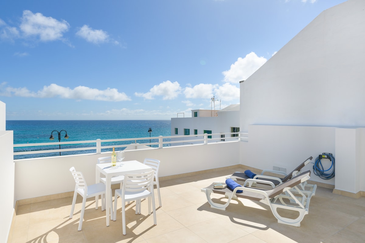 A balcony offers a stunning view of the ocean, featuring a table with chairs and two sun loungers. The space is brightened by natural light, and the sea can be seen in the background, complemented by a clear blue sky with scattered clouds.