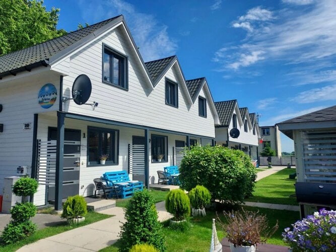 A row of two-story holiday homes showcases a light-colored exterior with a series of peaked roofs. Colorful blue outdoor seating is arranged in front of the homes, surrounded by well-maintained green shrubs and ornamental plants. Clear blue skies provide a bright backdrop.