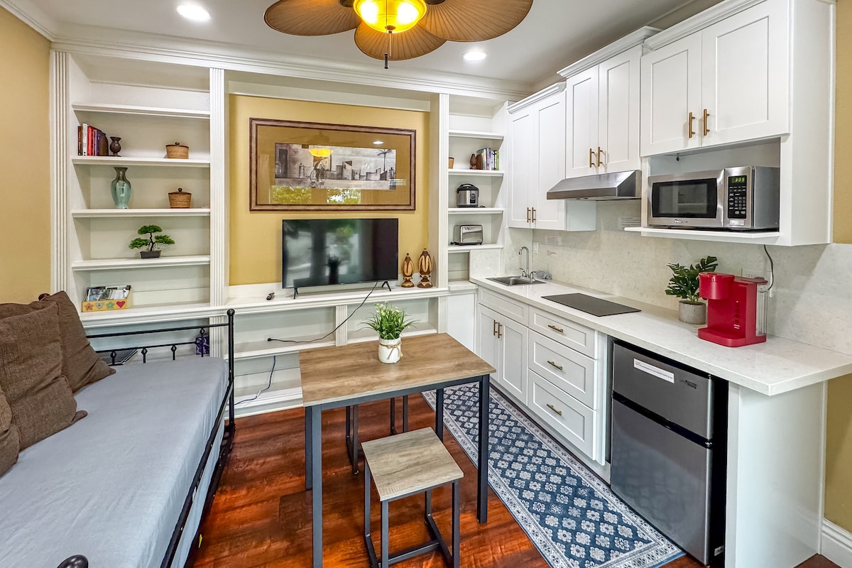 An inviting indoor living area presents a twin daybed, a dining table with two stools, and a well-equipped kitchenette containing a mini fridge, microwave, and Keurig coffee maker. Light wood cabinetry and decorative shelves enhance the space, complemented by a Smart TV and a potted plant.