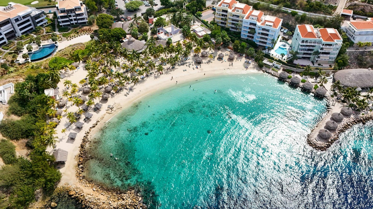 An aerial view captures a calm beach with gentle waves lapping at the shore. Umbrellas and lounge chairs are arranged on the sandy beach, surrounded by palm trees. Nearby, modern white buildings with red-roofed structures are visible, setting a serene tropical atmosphere.