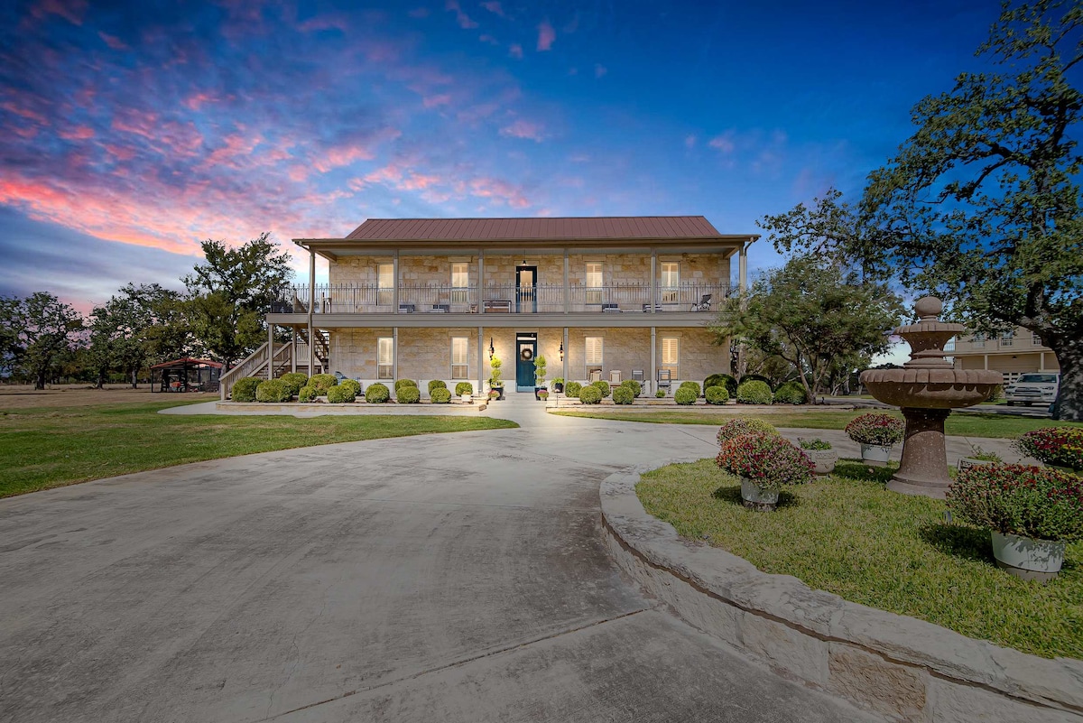 The exterior of a classic German-style estate is displayed, featuring a two-story structure with a welcoming front entrance. Neatly trimmed bushes surround the home, while a fountain adds a peaceful element. The driveway curves gently, leading to the expansive lawn under a colorful sunset.