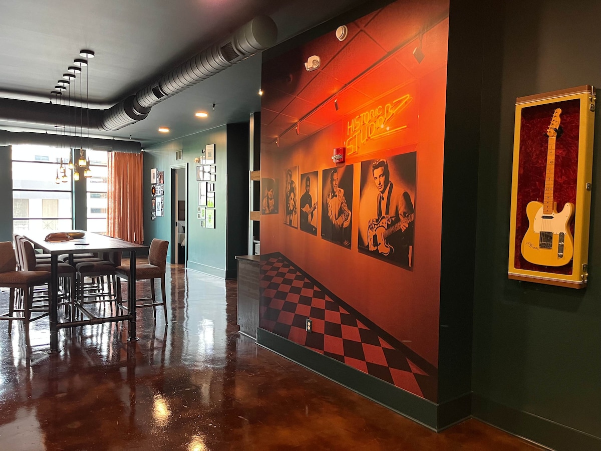 A vibrant dining area features a long table surrounded by high chairs. A wall is adorned with black and white photographs of musicians, complemented by a bright orange neon sign. The space boasts glossy concrete flooring and large windows that convey natural light.