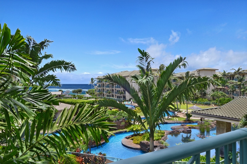 A scenic view from a private lanai showcases lush palm trees framing a vibrant lagoon-style pool. The ocean glistens in the distance under a clear blue sky, while the resort's tropical landscaping adds a serene ambiance to the setting.