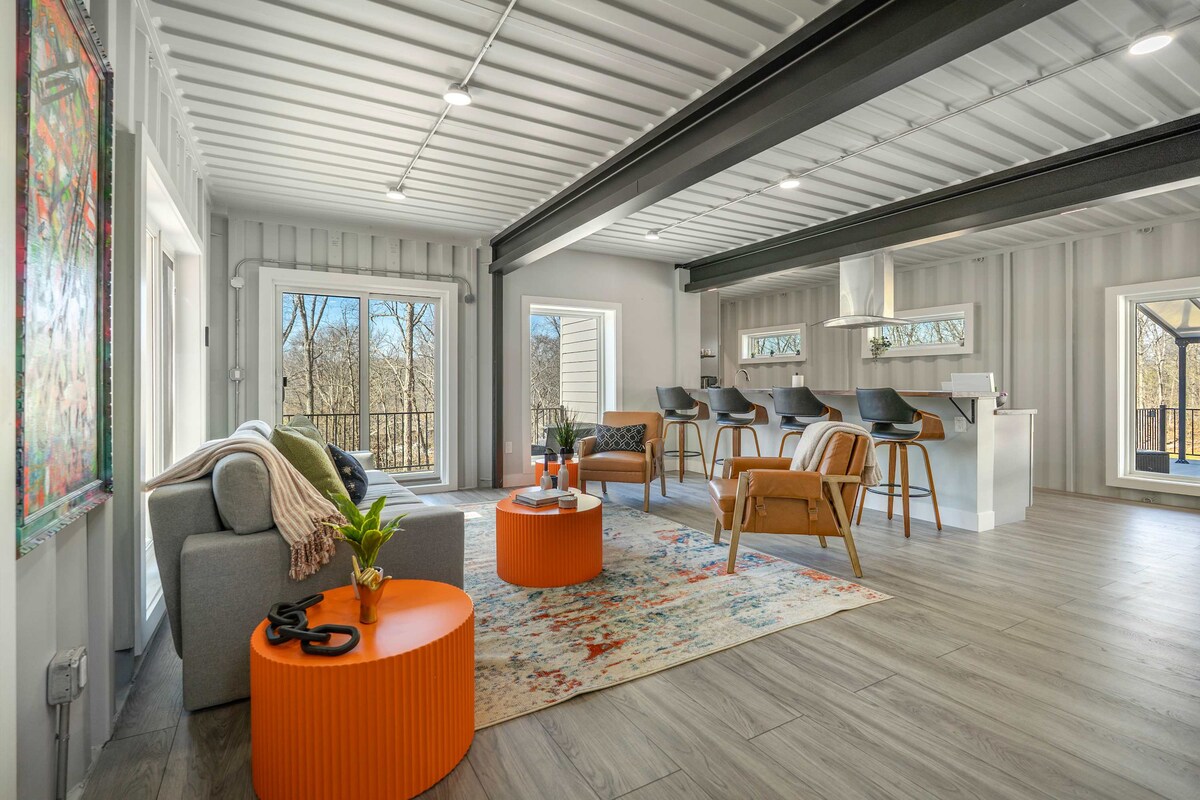 An open living space features large windows allowing natural light to illuminate the room. A cozy sofa is paired with an orange accent table, and the area is adorned with a colorful rug. Bar stools line the kitchen counter in the background.
