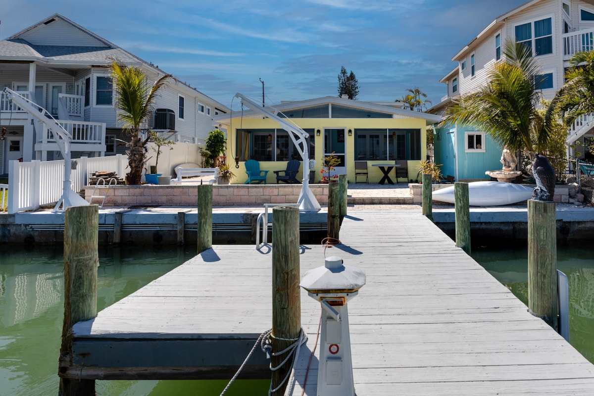 A wooden dock extends over calm water, leading to a colorful waterfront apartment. The building features large windows and a screened porch. Palm trees add greenery to the surroundings, while neighboring properties are visible on either side, contributing to a relaxed coastal atmosphere.