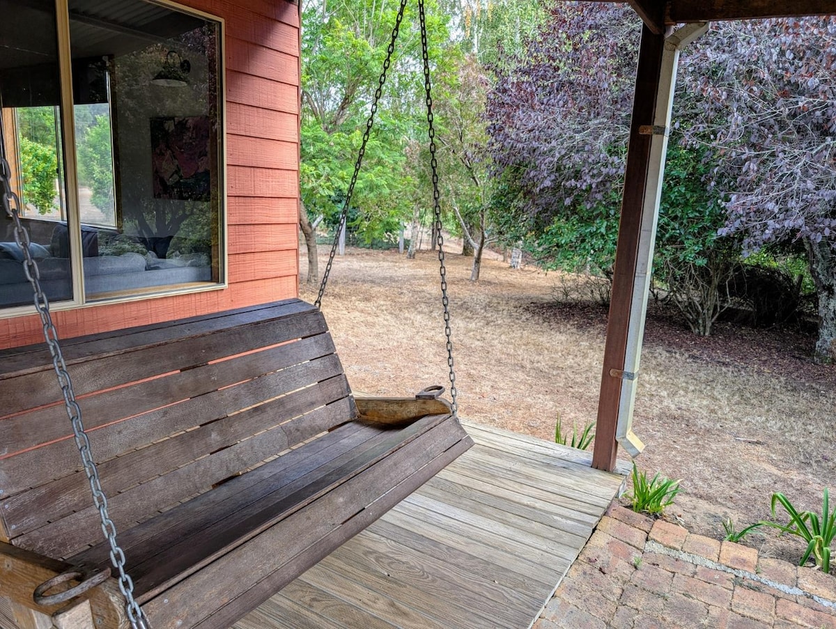 An outdoor swing seat is positioned on a wooden deck, offering a spot to relax. The background features a grassy area with trees and shrubs. The natural setting provides a serene atmosphere for enjoyment.