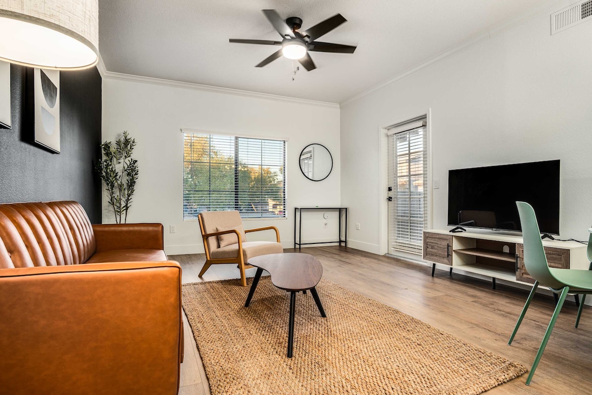 An inviting living area features a stylish brown leather sofa and a modern lounge chair. A round table stands in the center, surrounded by warm wood flooring. Large windows allow natural light to fill the space, complemented by a ceiling fan and wall-mounted television.