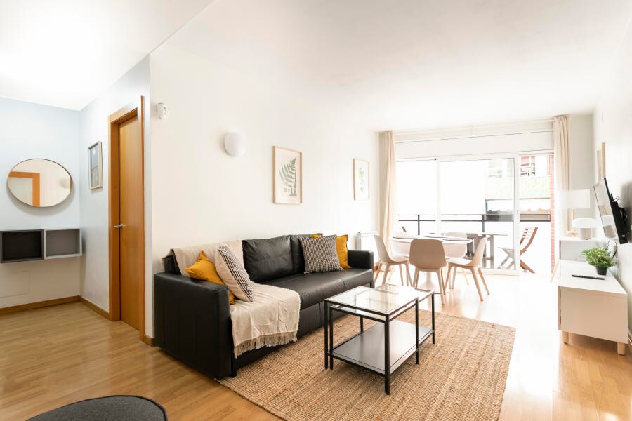 The living room features a comfortable black sofa adorned with decorative cushions and a light throw. A glass coffee table sits atop a natural fiber rug, complemented by a flat-screen TV and a small dining area with wooden chairs. Large windows allow natural light to fill the space.