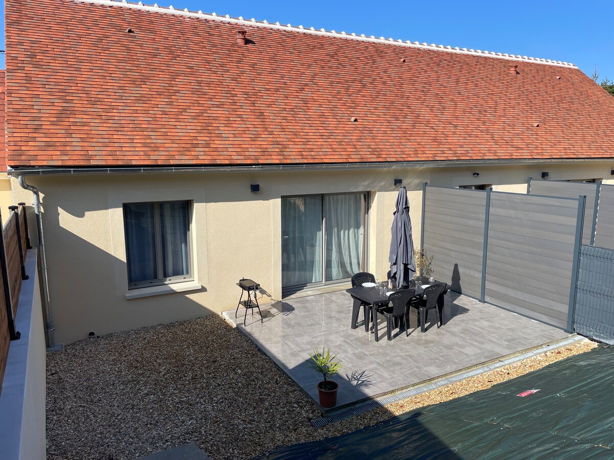 An outdoor terrace space is featured, furnished with a table and chairs, alongside an umbrella for shade. A small table and a potted plant are also visible. The surrounding area includes gravel and an enclosed fence, while the house's exterior is complete with a sloped roof.