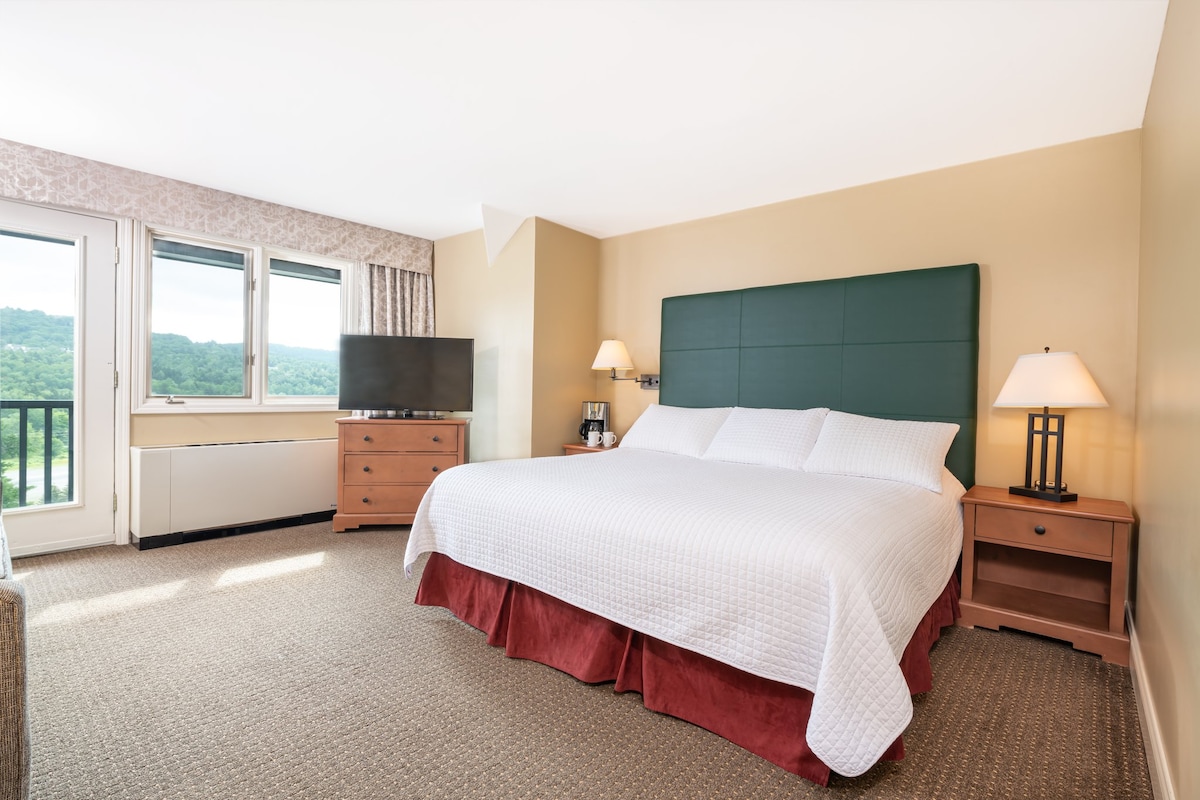 A spacious room features a comfortable king bed with crisp white linens. A television is placed on a wooden dresser. Two large windows allow natural light to fill the space, offering scenic views of the surrounding landscape. Soft lighting is provided by bedside lamps.