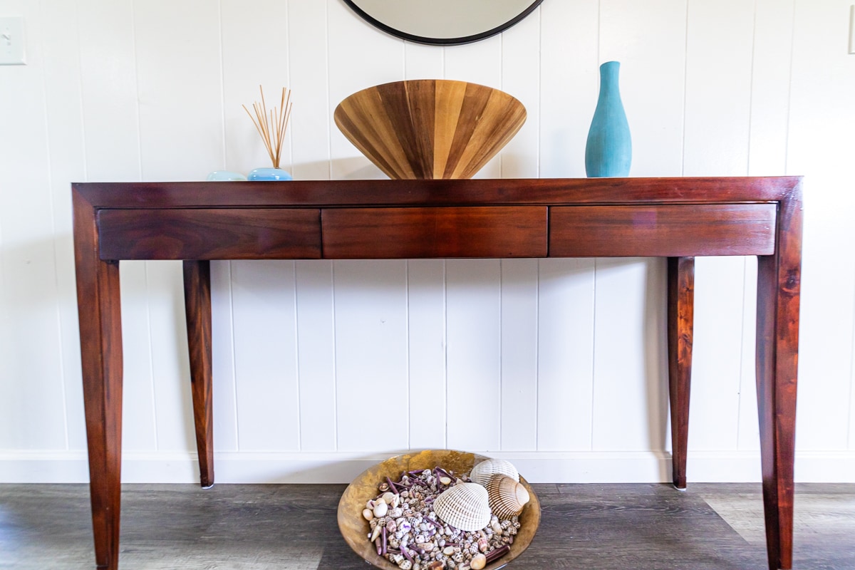 A wooden console table with a rich finish features three drawers and stands on slender legs. Decorative pieces include a large wooden bowl, a turquoise vase, and items arranged in a bowl at the base, showcasing an assortment of seashells.