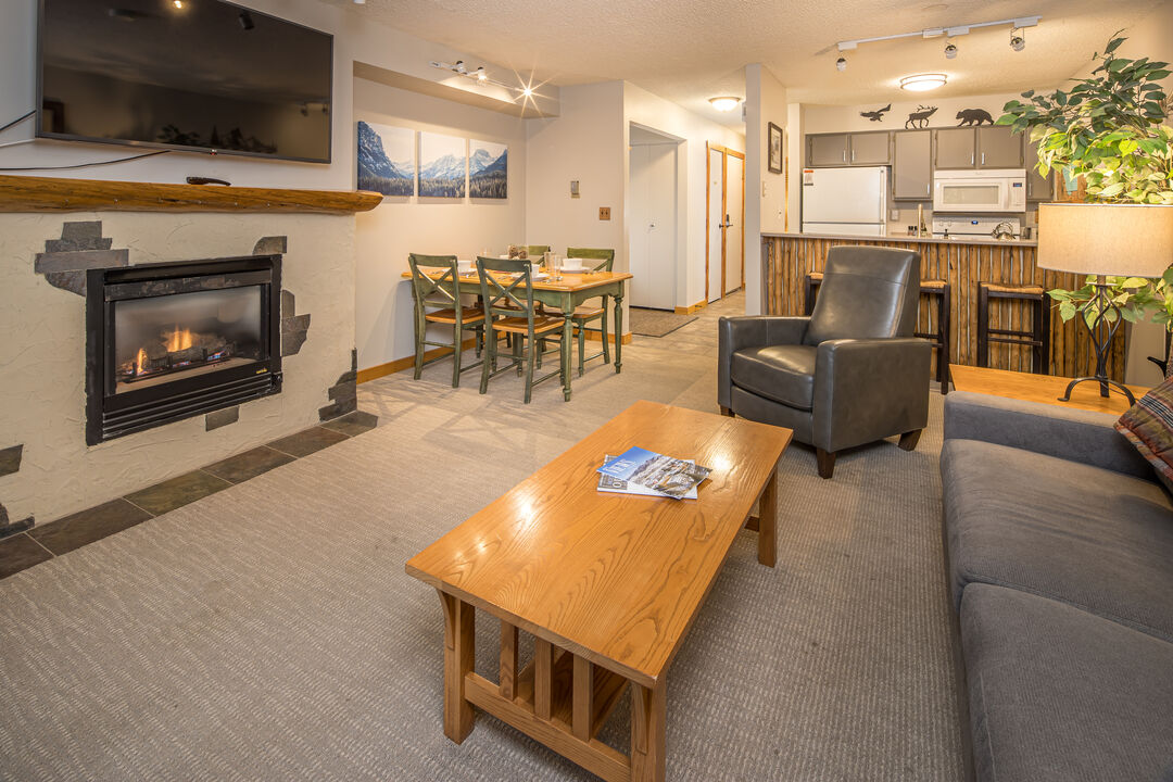 The open-concept living area features a comfortable seating arrangement centered around a fireplace. A wooden coffee table occupies the center, while a dining table with green chairs is visible in the background. The kitchen is equipped with modern appliances and offers a warm, welcoming ambiance.