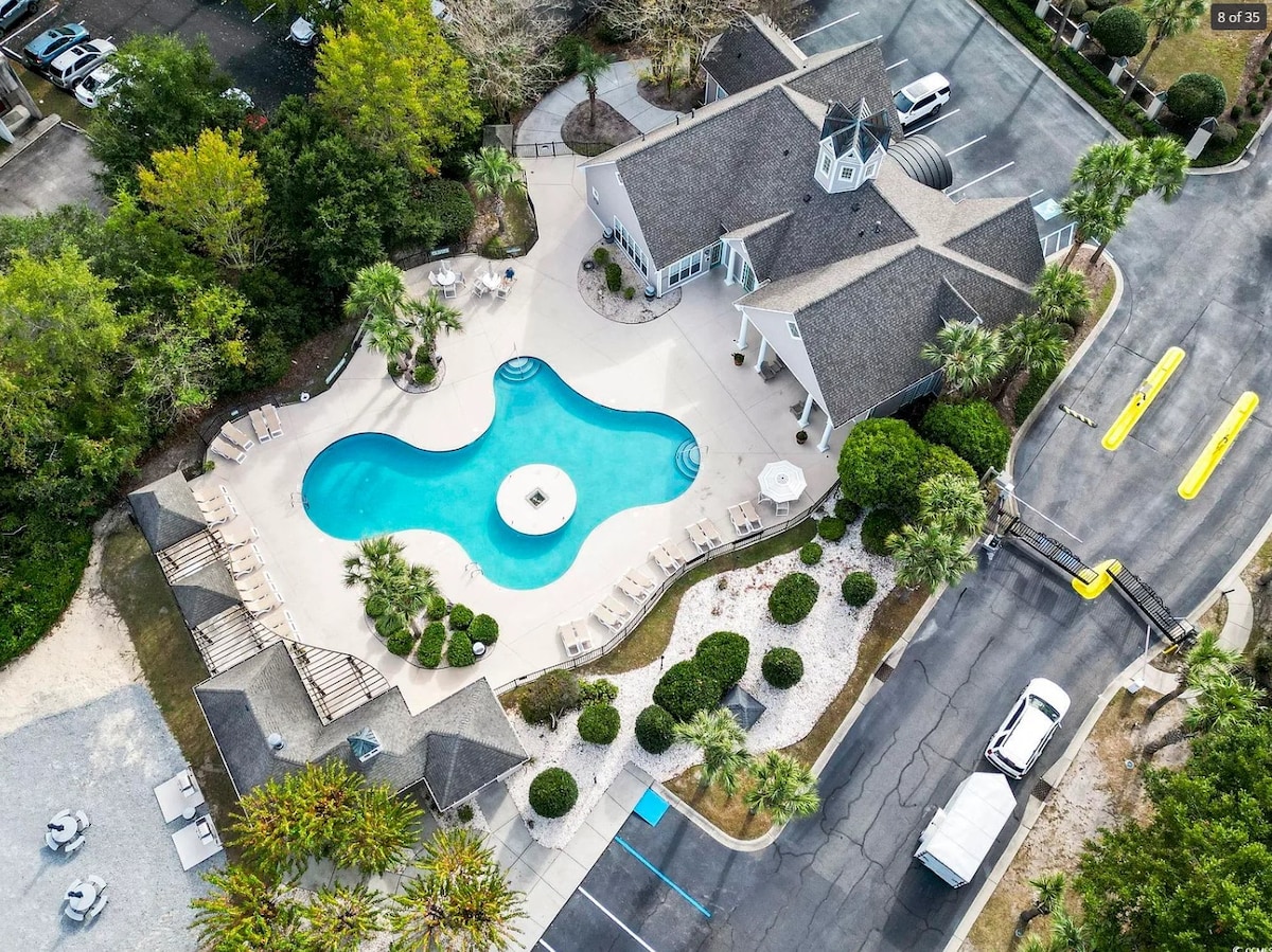 An aerial view showcases the outdoor pool area of the community, surrounded by landscaped gardens and palm trees. The pool features a curved design with shaded lounge chairs positioned nearby. The clubhouse is visible, emphasizing the welcoming environment of the complex.