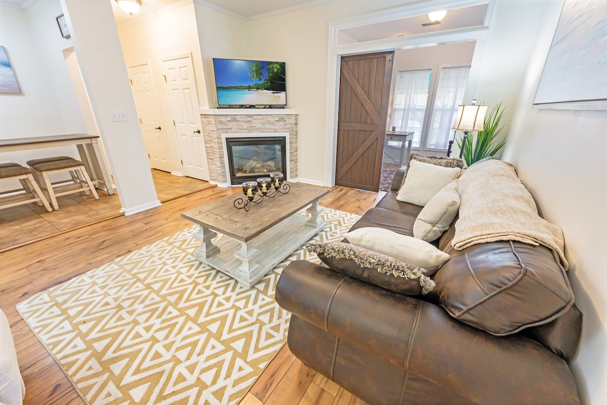 A cozy living room is depicted, featuring a comfortable sectional sofa adorned with pillows. A coffee table rests on a patterned rug, and a fireplace is placed beneath a wall-mounted television. Natural light enters through nearby windows, complementing the warm wood flooring.