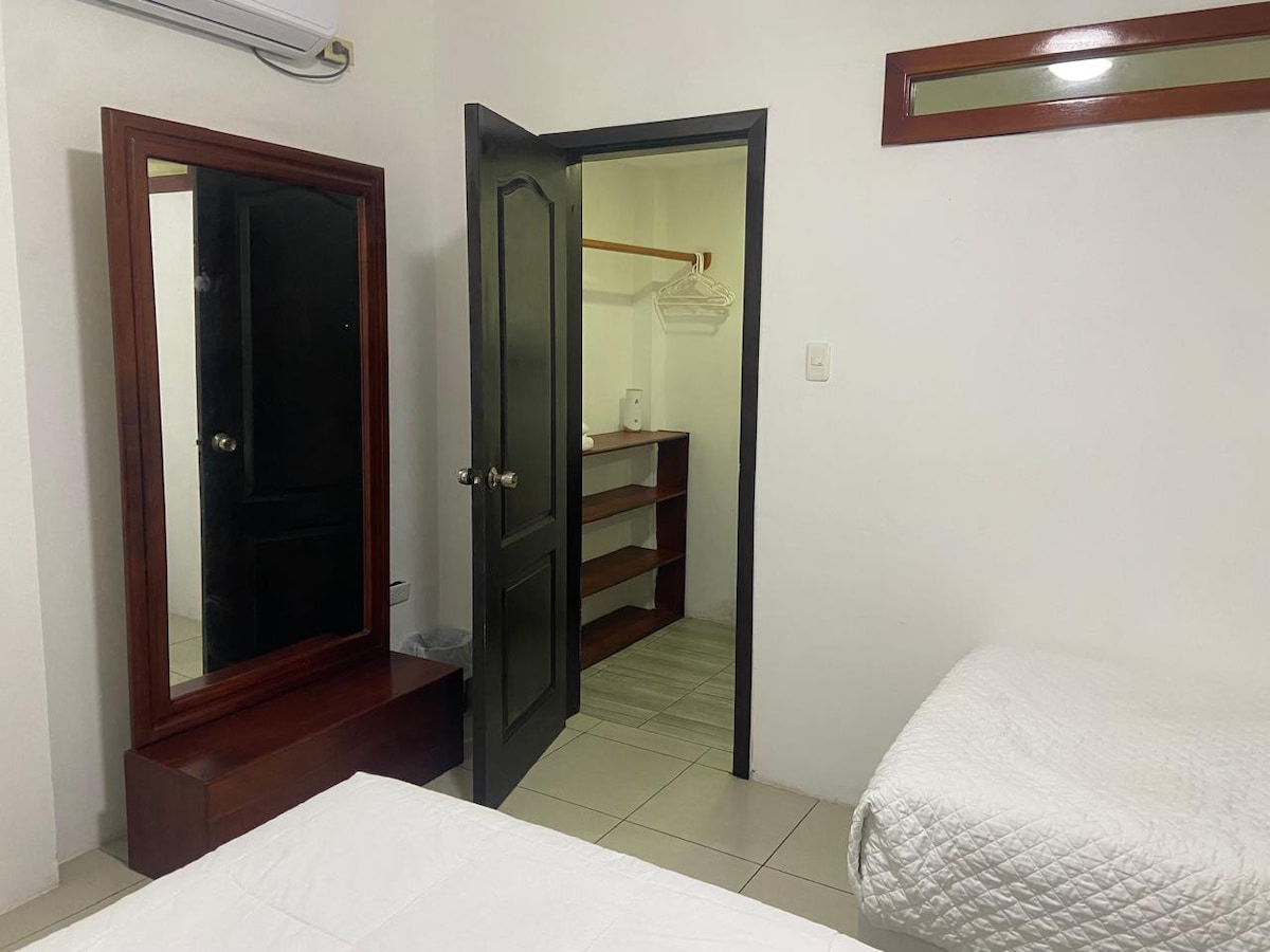 An interior view of a cozy guest room is shown, featuring two beds with white covers. A full-length mirror is positioned near the door, and a closet space with shelves is visible through an open door. Light-colored tile flooring enhances the room's spaciousness.