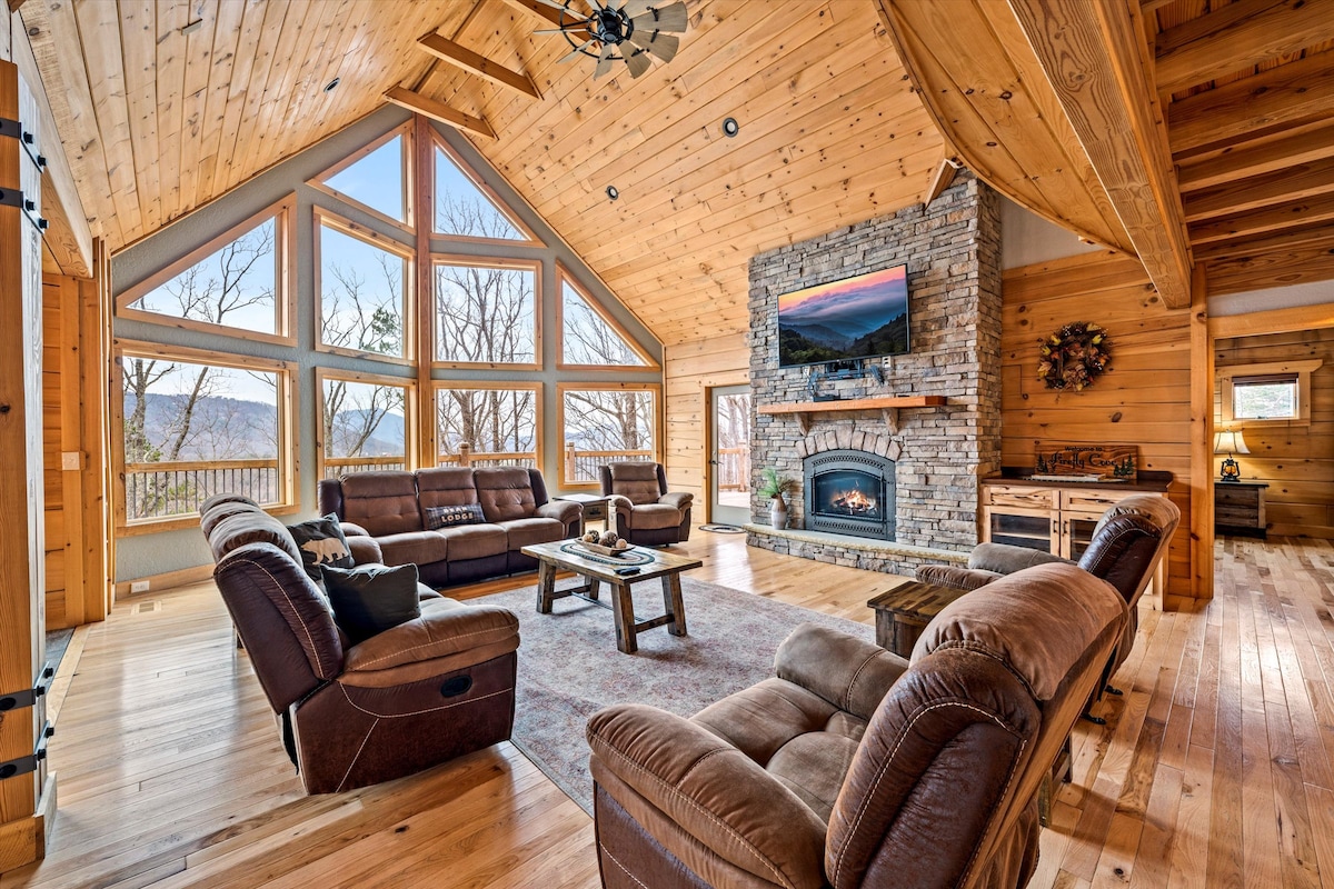 A spacious living area features vaulted wooden ceilings with large floor-to-ceiling windows, offering panoramic views of the mountains. Comfortable brown seating surrounds a central coffee table, with a stone fireplace and a mounted television creating a cozy family gathering space.