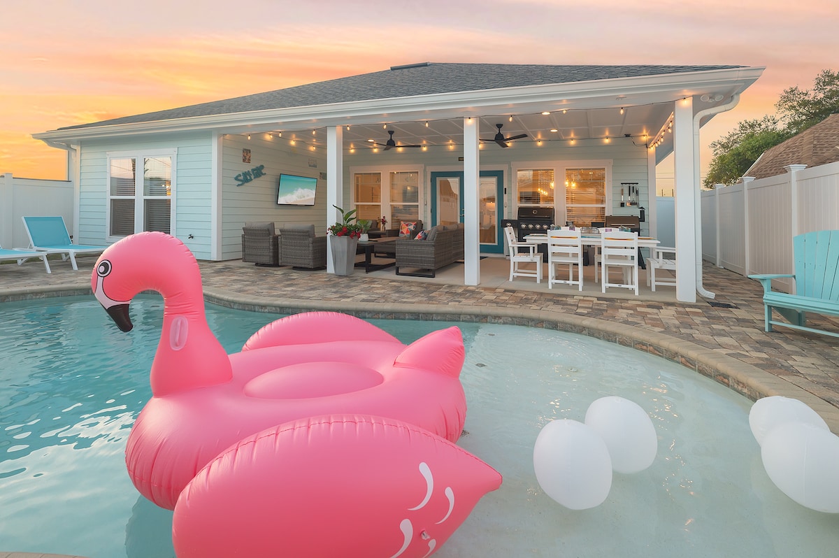 A pink inflatable flamingo floats in the inviting pool, reflecting the warm hues of the sunset. In the background, a cozy covered patio is furnished with comfortable seating and a dining area. Strings of lights add a subtle glow, enhancing the serene outdoor space.