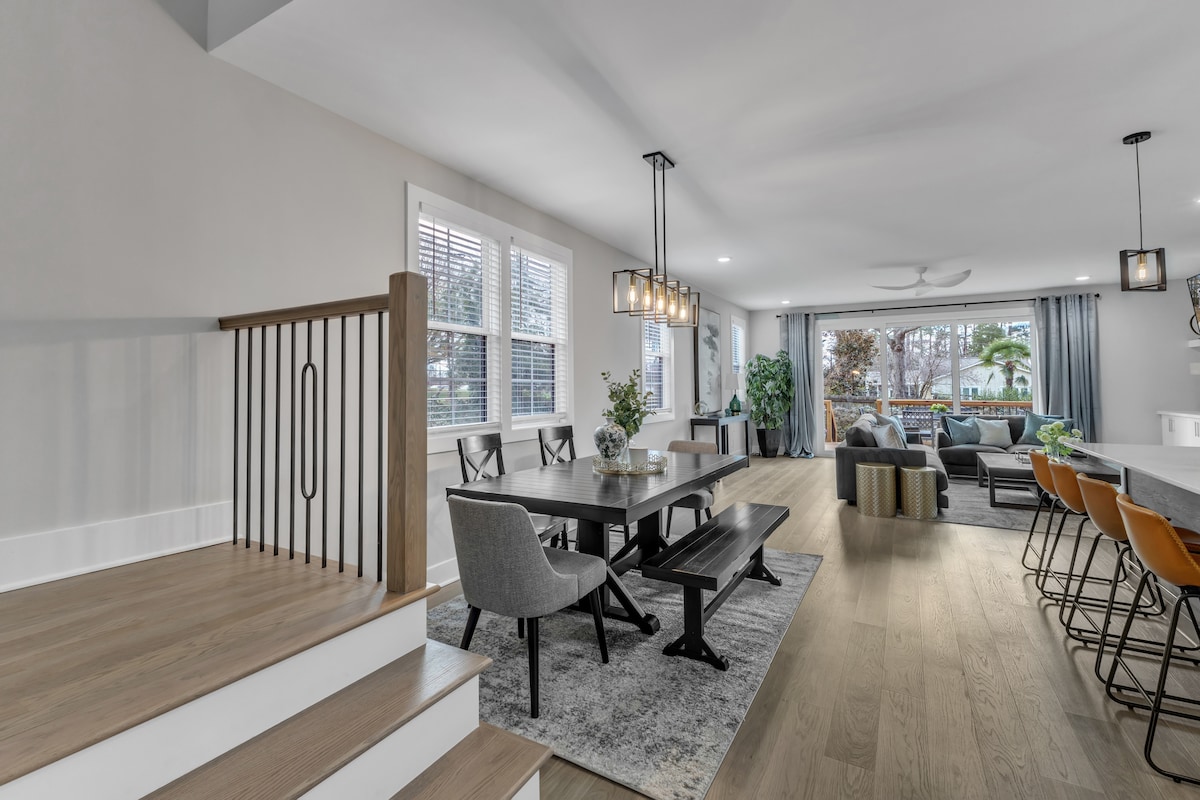 An open dining area is furnished with a large table surrounded by chairs, while a cozy living space is visible in the background. Natural light pours in through large windows, highlighting a neutral color palette and modern decor elements throughout the space.