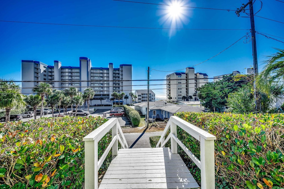 A scenic pathway leads through lush greenery toward a view of nearby beachside buildings. Bright sunlight illuminates the clear blue sky, while palm trees frame the peaceful landscape. The beach is accessible just a short walk away, enhancing the inviting atmosphere.