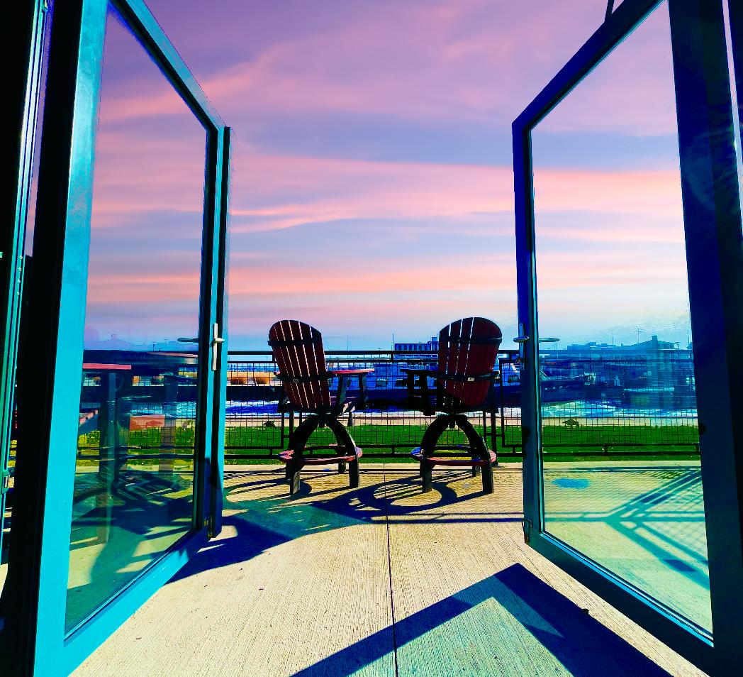 A private balcony is shown through open glass doors, featuring two wooden chairs and a small table. The sky is painted in soft hues of pink and purple, creating a serene outdoor space overlooking the landscape.