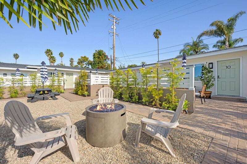 A communal outdoor area features a fire pit surrounded by light-colored Adirondack chairs. The space is enhanced by tropical plants and palm trees, with picnic tables available in the distance. The buildings are modern and painted in a soft palette, creating a welcoming environment.