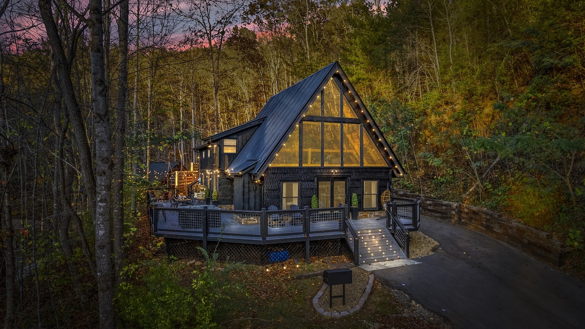The modern log cabin, set against a backdrop of trees, features large glass windows that reflect the warm tones of sunset. A spacious deck invites outdoor gatherings, while lights accentuate the architecture, creating an inviting presence in the secluded mountain setting.