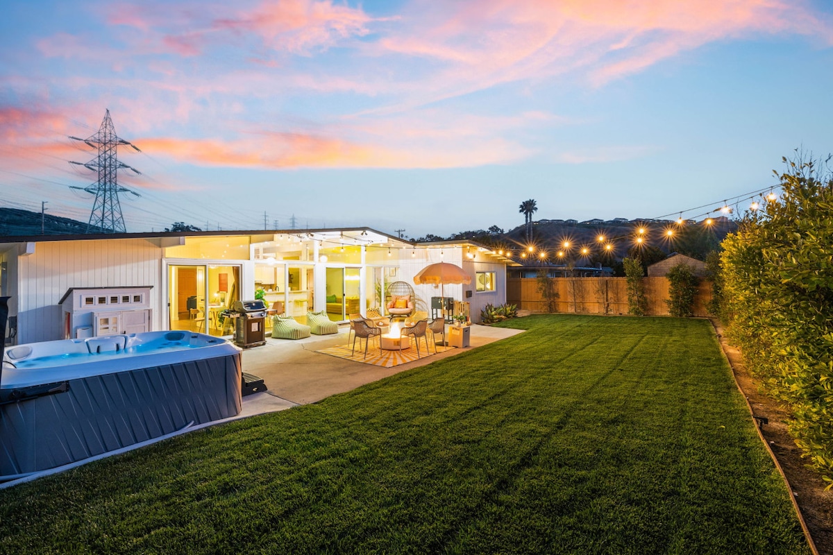 The backyard is illuminated by soft lights, showcasing a spacious lawn and relaxing hot tub. Outdoor seating areas are arranged around a fire pit, and a large umbrella provides shade. The colorful sunset reflects off the modern home, blending indoor and outdoor spaces.