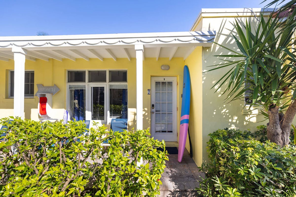 The exterior of a bright yellow studio apartment is shown, featuring a covered patio with white columns. A colorful surfboard leans against the wall next to the entrance, framed by lush greenery and potted plants, creating a welcoming outdoor space.