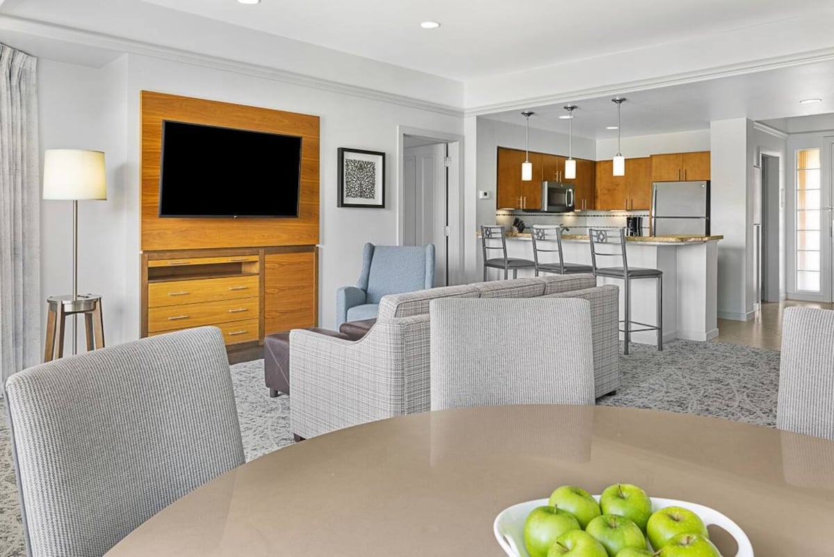 An open living area features a round dining table with green apples as a centerpiece. Comfortable chairs and a sofa are positioned for relaxation, while a flat-screen TV is mounted on a wooden cabinet. A full kitchen is visible in the background.