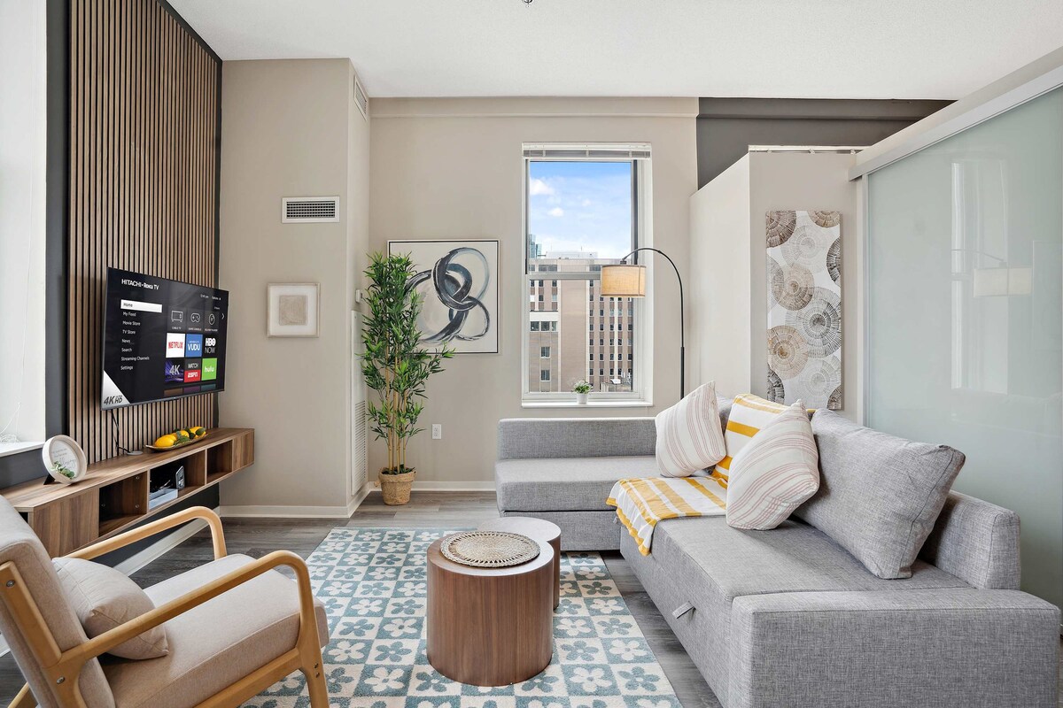 The living room features a modern design with a comfortable sofa and accent chairs. A 55" Smart TV is mounted on the wall, with a stylish coffee table in front. Natural light enters through the window, highlighting the soft textures of the area rug.