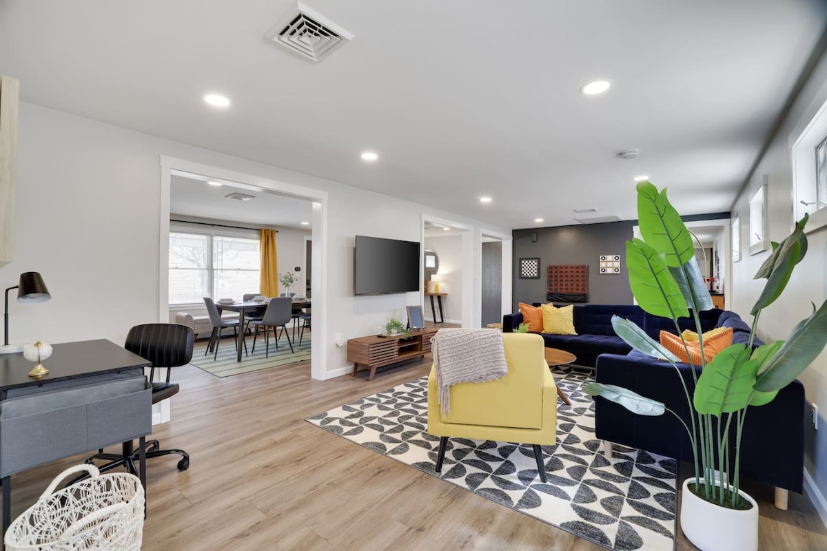 A spacious living area features a mix of comfortable seating, including a navy sofa and a yellow armchair. A modern coffee table rests on a patterned rug, while large windows allow natural light to brighten the room. A smart TV is mounted on the wall opposite a sleek work desk.