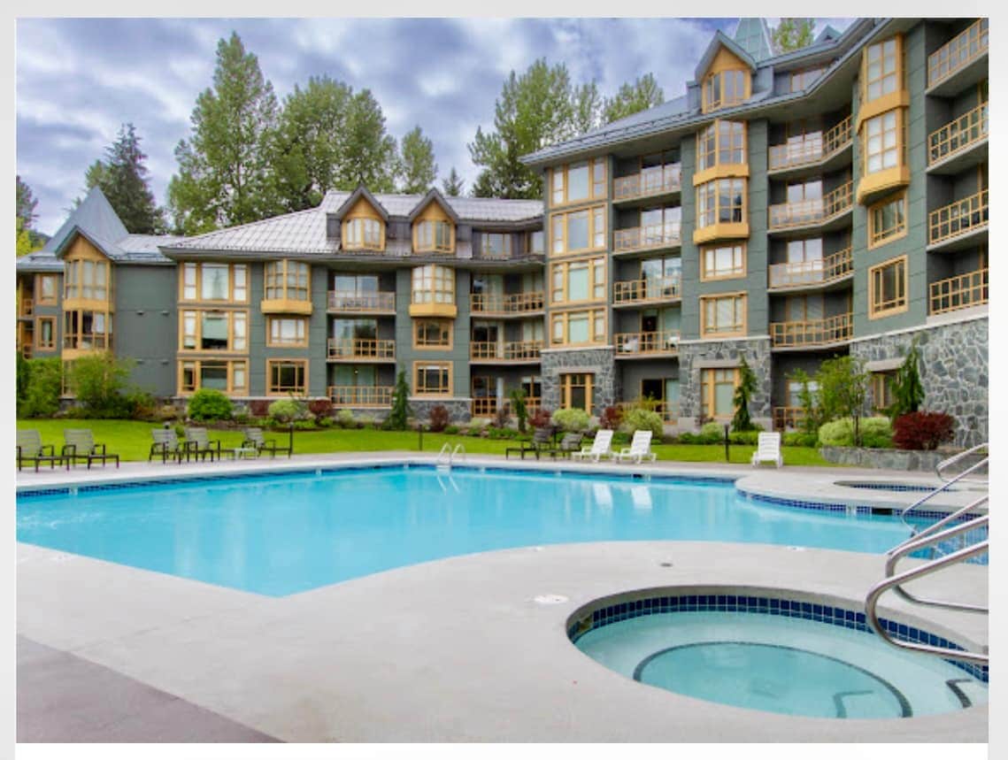 The outdoor area features a large swimming pool with a hot tub nearby, surrounded by neatly arranged lounge chairs. The modern building showcases expansive windows, offering views of the lush greenery and a well-maintained landscape in the backdrop.