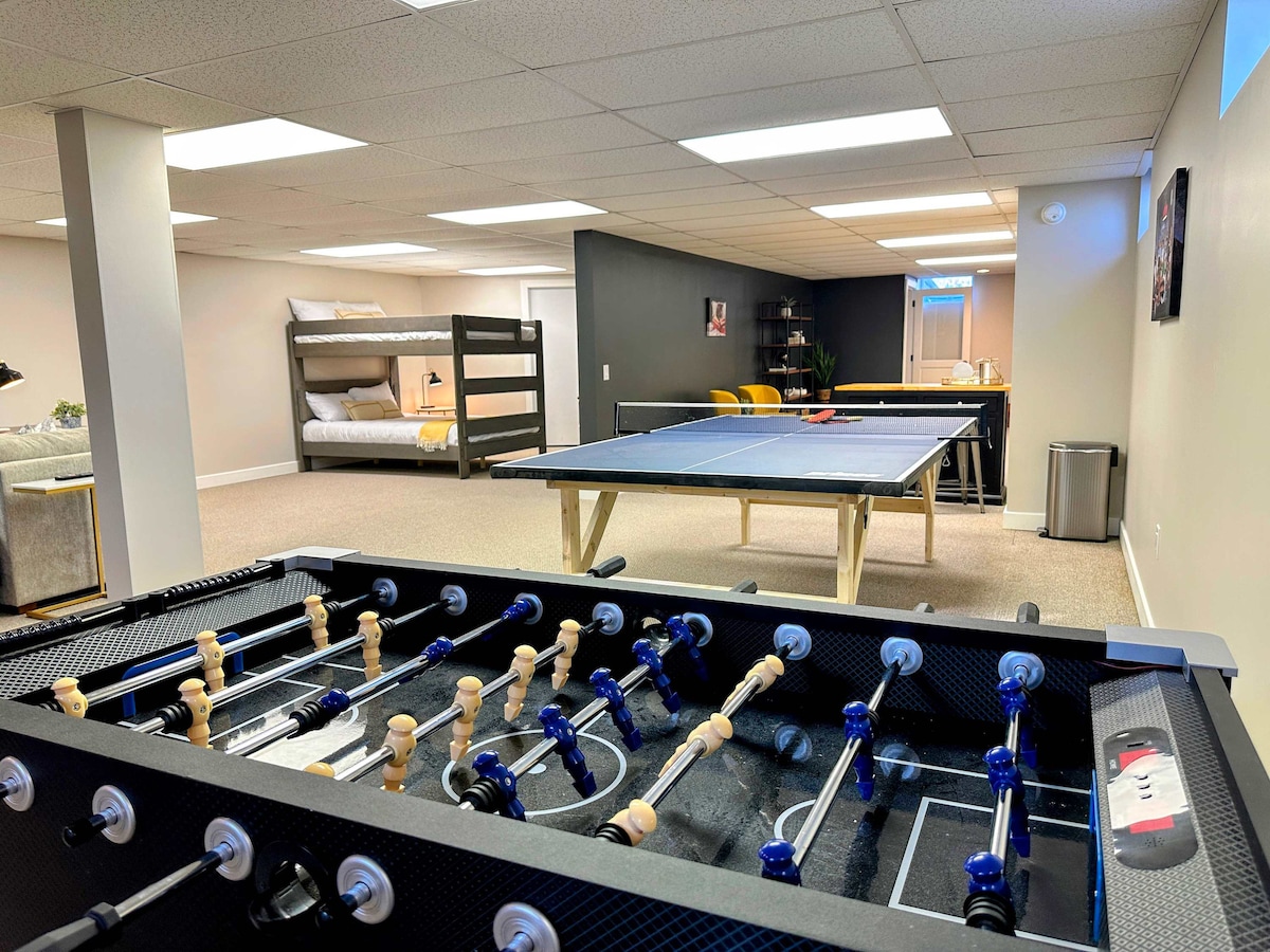 The spacious game room features a foosball table in the foreground and a ping pong table in the background. A cozy bunk bed is visible to the side, along with soft lighting and neutral-colored walls, creating an inviting space for recreation and relaxation.