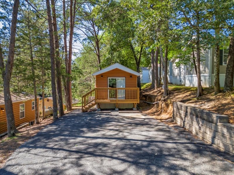 A charming park model home is featured, nestled among trees at Gateway RV Campground. A wooden deck with a railing provides access to the entrance, while the surrounding environment creates a serene and natural atmosphere. A paved area offers convenient parking for guests.