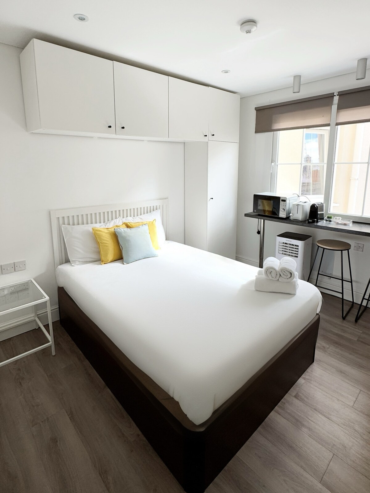 The spacious sleeping area features a bed with cozy linens and decorative pillows. A minimalistic side table is positioned alongside. Natural light floods the room through large windows, which include beige shades. A compact kitchenette with essential appliances is visible in the corner.