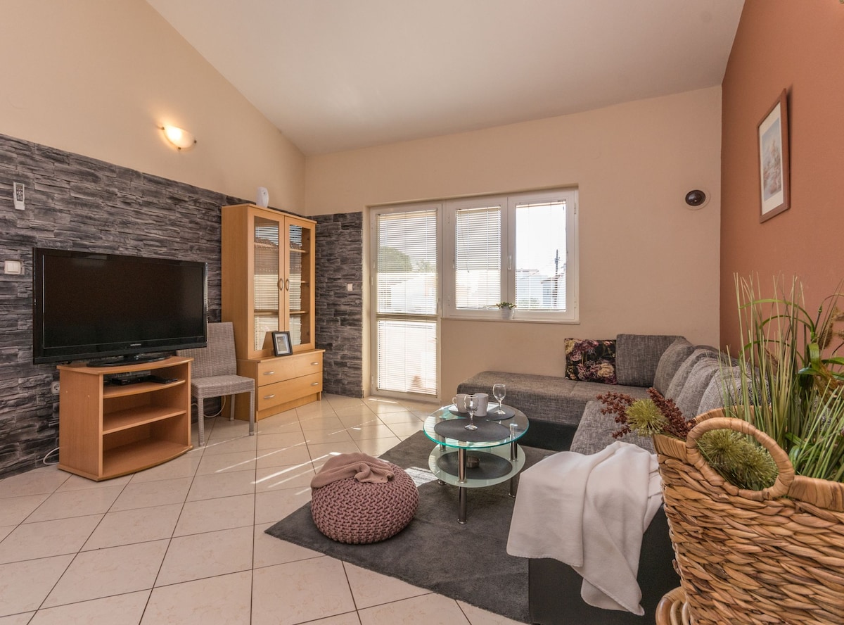 A living area features a comfortable gray sectional sofa, a glass coffee table, and a wooden entertainment unit with a television. Natural light filters through large windows, while decorative plants and a soft rug add warmth to the space.