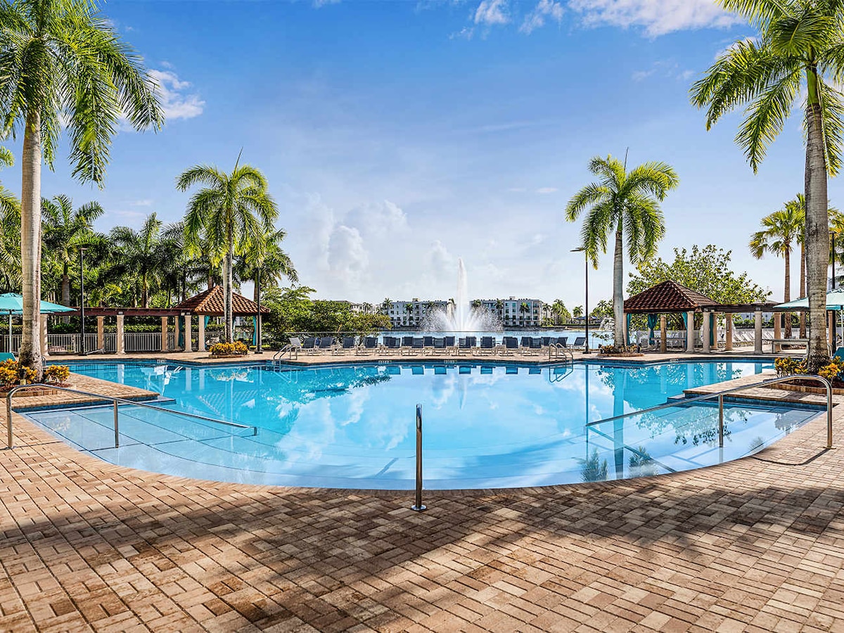 A large, inviting swimming pool is surrounded by well-manicured landscaping, featuring palm trees and vibrant flowers. Lounge chairs line the poolside, and a fountain is visible in the background, adding a serene element to the setting.