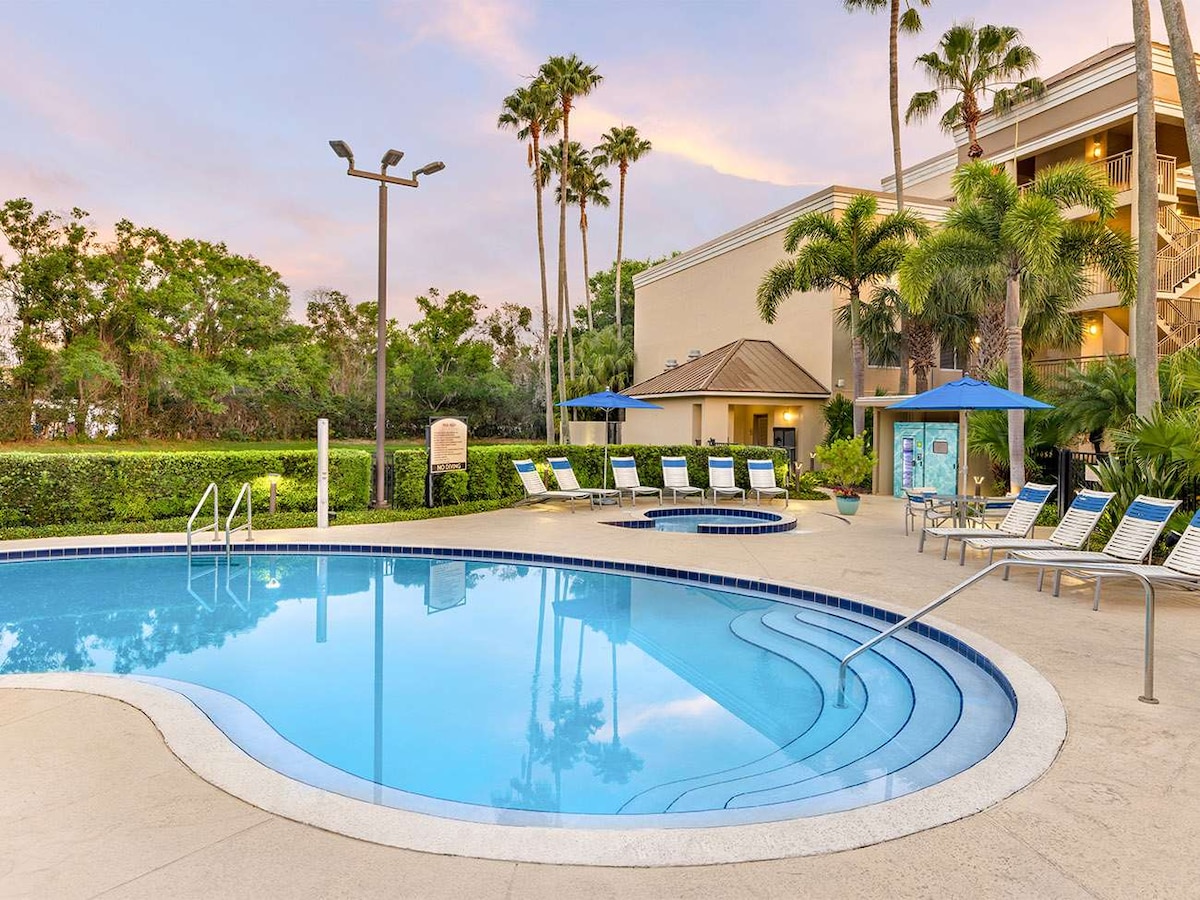 The outdoor swimming pool is surrounded by palm trees and features lounge chairs for relaxation. A hot tub is adjacent to the pool, and a shade structure provides relief from the sun. The serene landscape is complemented by the soft glow of evening light.