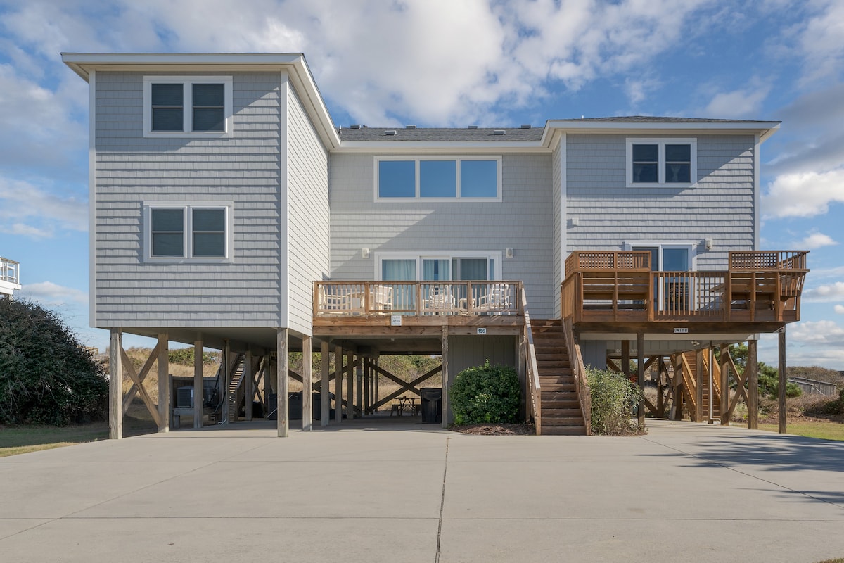 The exterior of Coop's Cottage features a two-story structure elevated on pilings, showcasing multiple windows that provide natural light. A spacious deck is accessible from the second level, complemented by a staircase leading to the ground level. The surrounding area includes landscaped greenery.