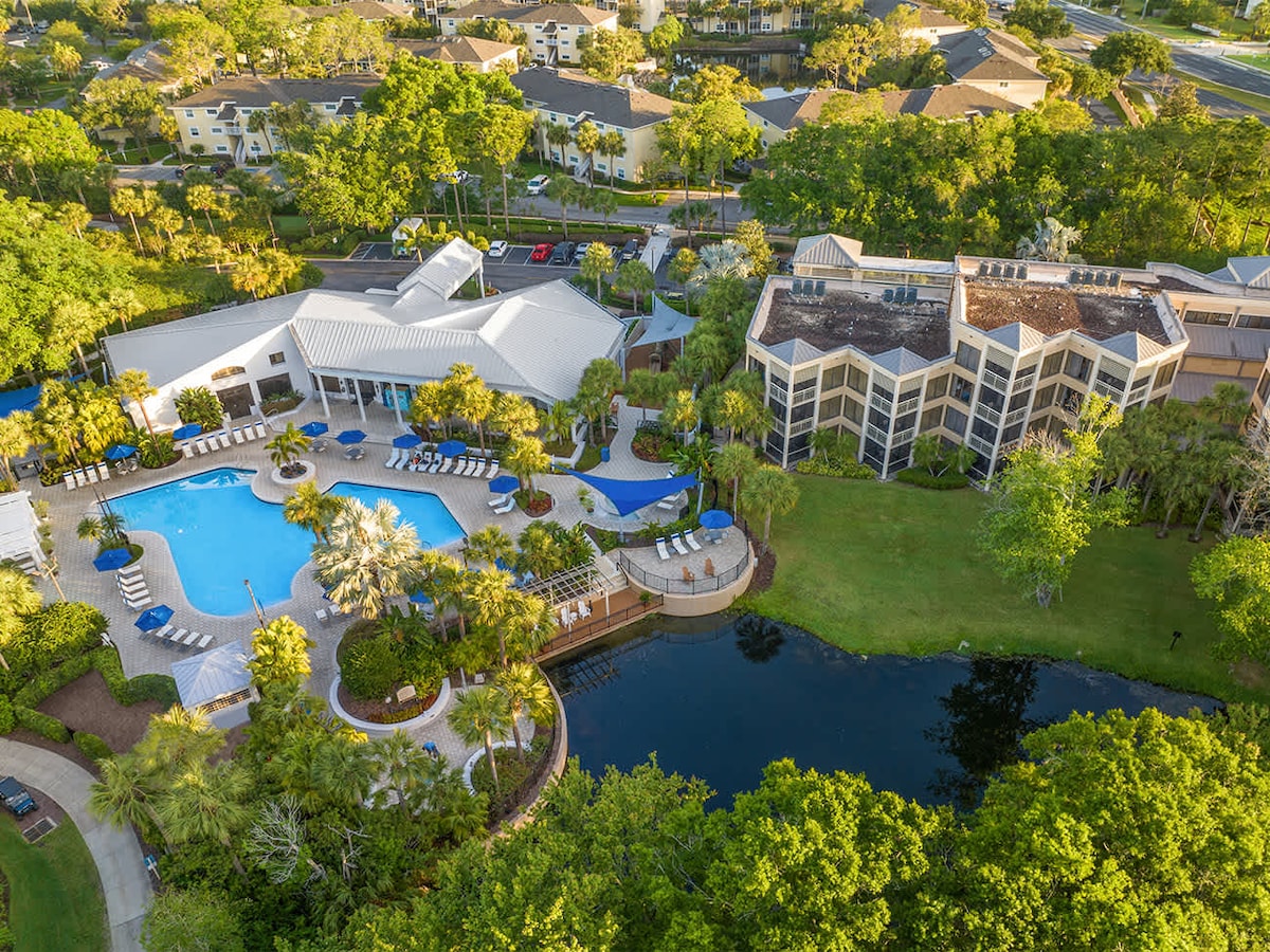 An aerial view showcases the resort's outdoor pool area surrounded by palm trees and lounge chairs. The landscaped grounds feature a serene pond, with multiple buildings visible in the background, harmoniously integrated into the lush natural setting.