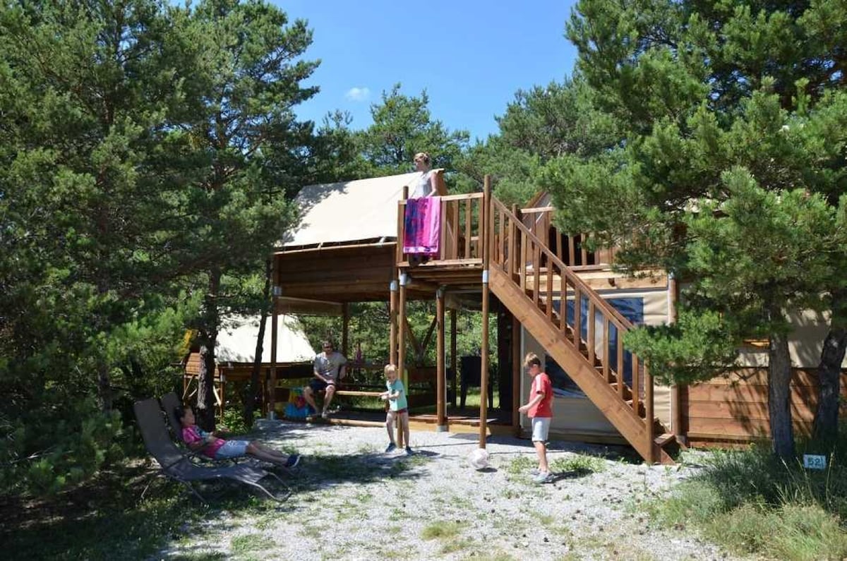 A wooden chalet is positioned among greenery, featuring a raised deck accessed by stairs. Children are seen playing in the outdoor space, while one child stands on the deck, holding a towel. Nearby, a lounge chair is visible, blending with the natural surroundings.