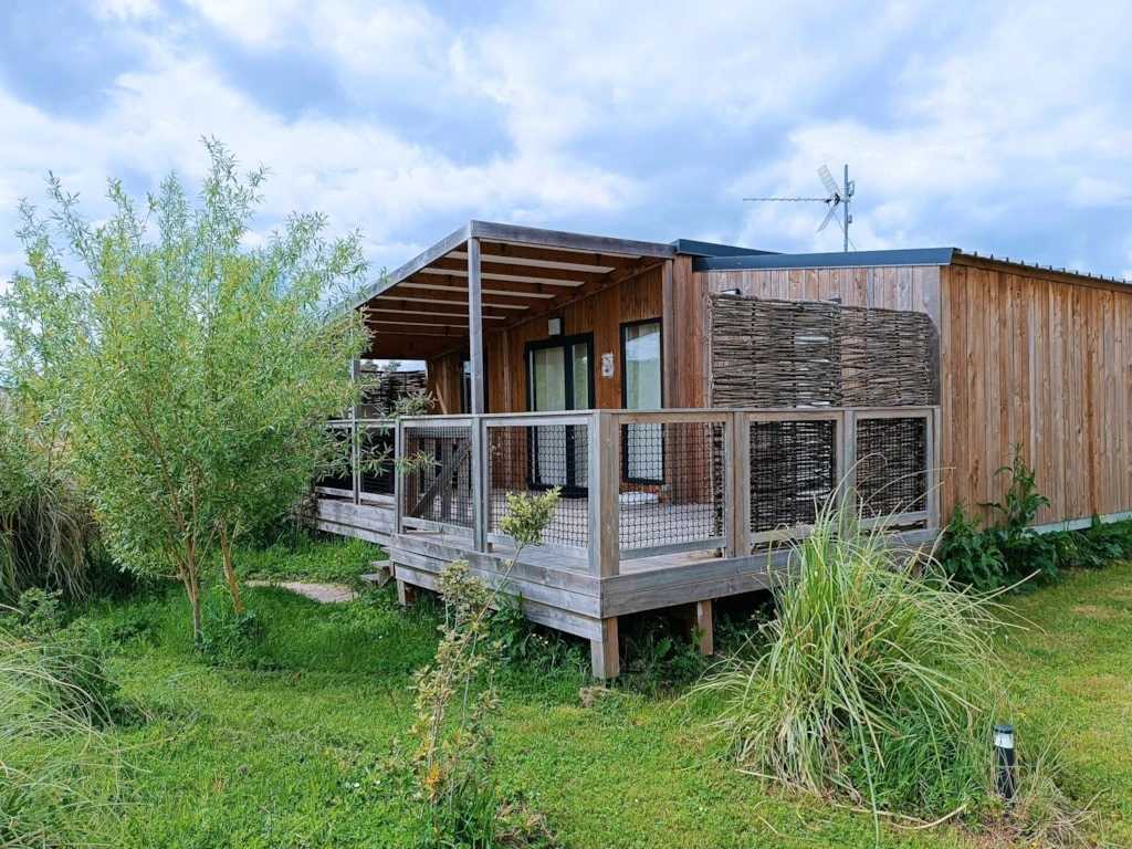 A modern wooden structure is surrounded by lush greenery, featuring a spacious deck with a simple railing. Large glass doors provide access to indoor areas. Nearby foliage adds a natural element, while the sky reflects a mix of clouds.